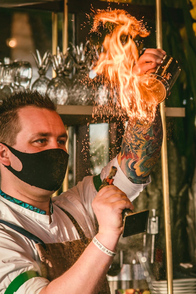 A bartender wearing a mask skillfully creates a fire show with cocktail preparation, adding flair at a bar.