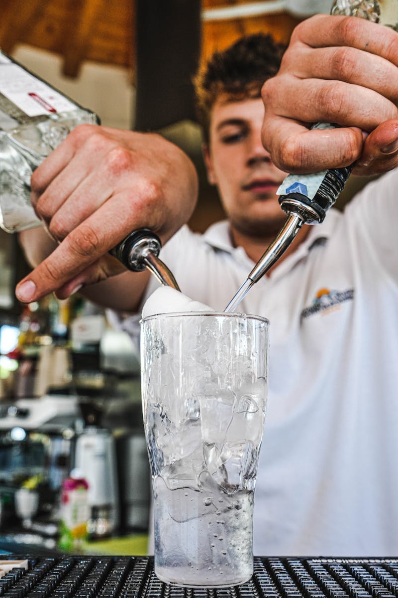 A bartender skillfully pours two alcoholic drinks over ice at a bar, showcasing a lively atmosphere.