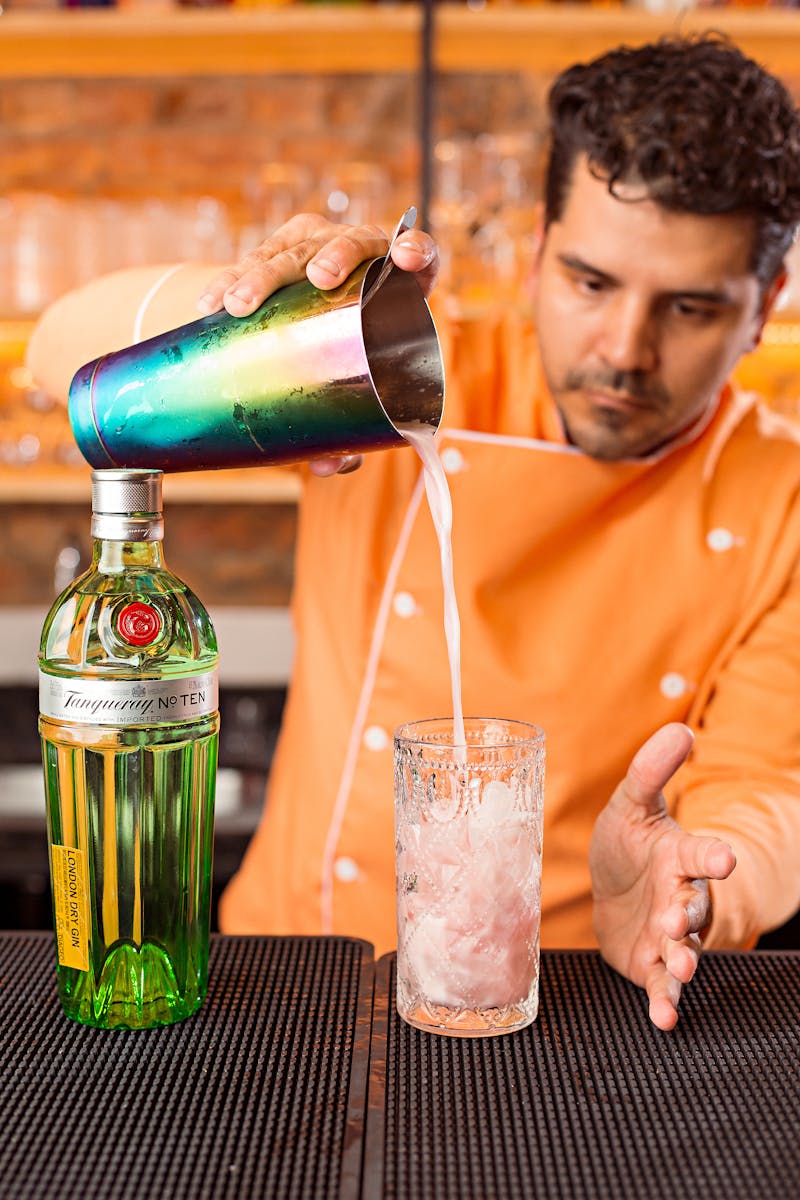 A skilled bartender pours gin into a cocktail glass with ice at a vibrant bar setting.